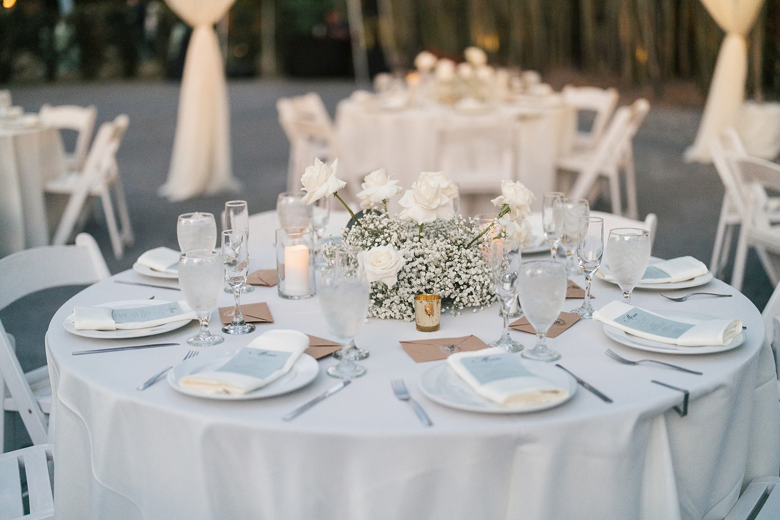 Round reception table set with white roses, baby's-breath, candles, menu cards, and kraft envelopes at dusk, an example of the detail-first frames that open a gallery