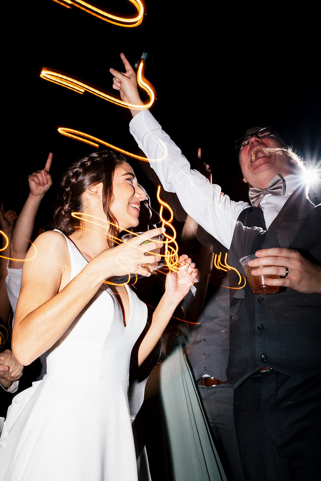 Bride laughing and dancing with a guest on the reception dance floor, streaks of warm light trailing through the dark night sky above them.