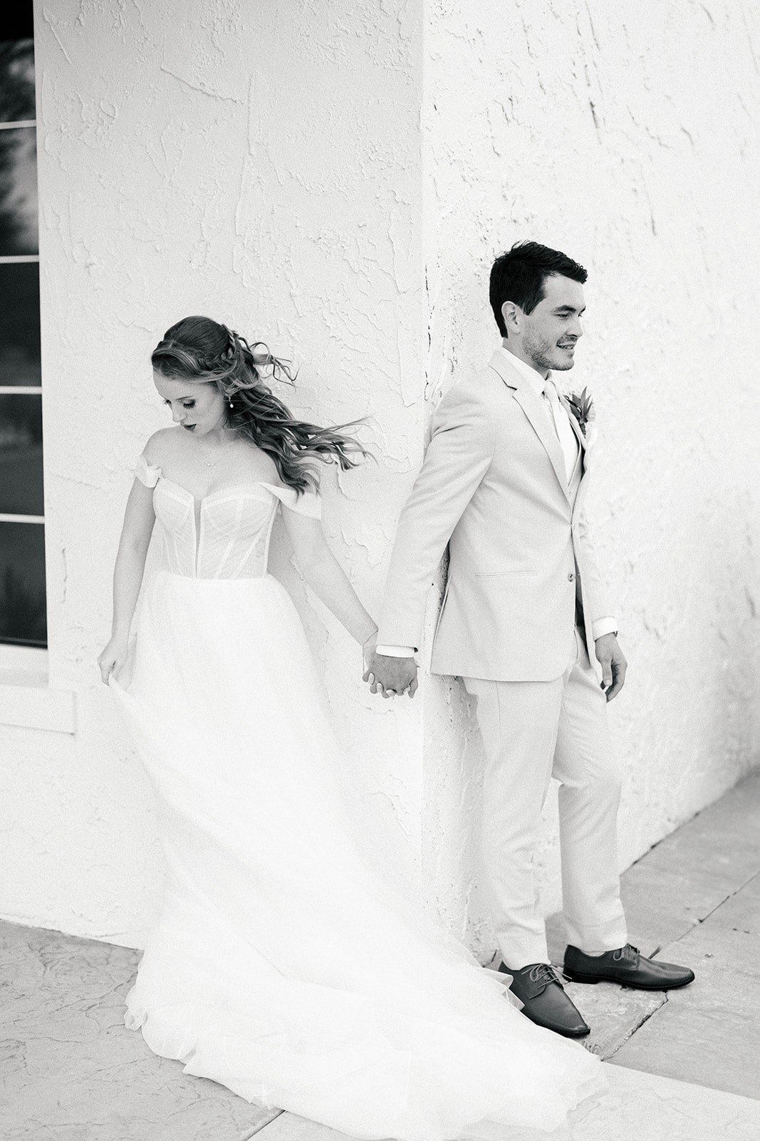 Bride looks down as wind blows through her hair while holding hands with the groom, both leaning against a stucco wall, black and white portrait.