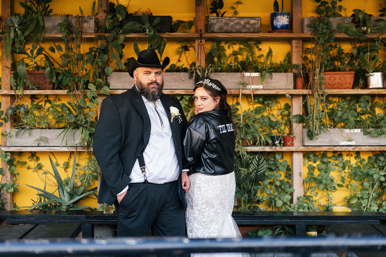 Sydney in a 'Til Death leather jacket beside Manny in a cowboy hat before a plant-lined yellow wall