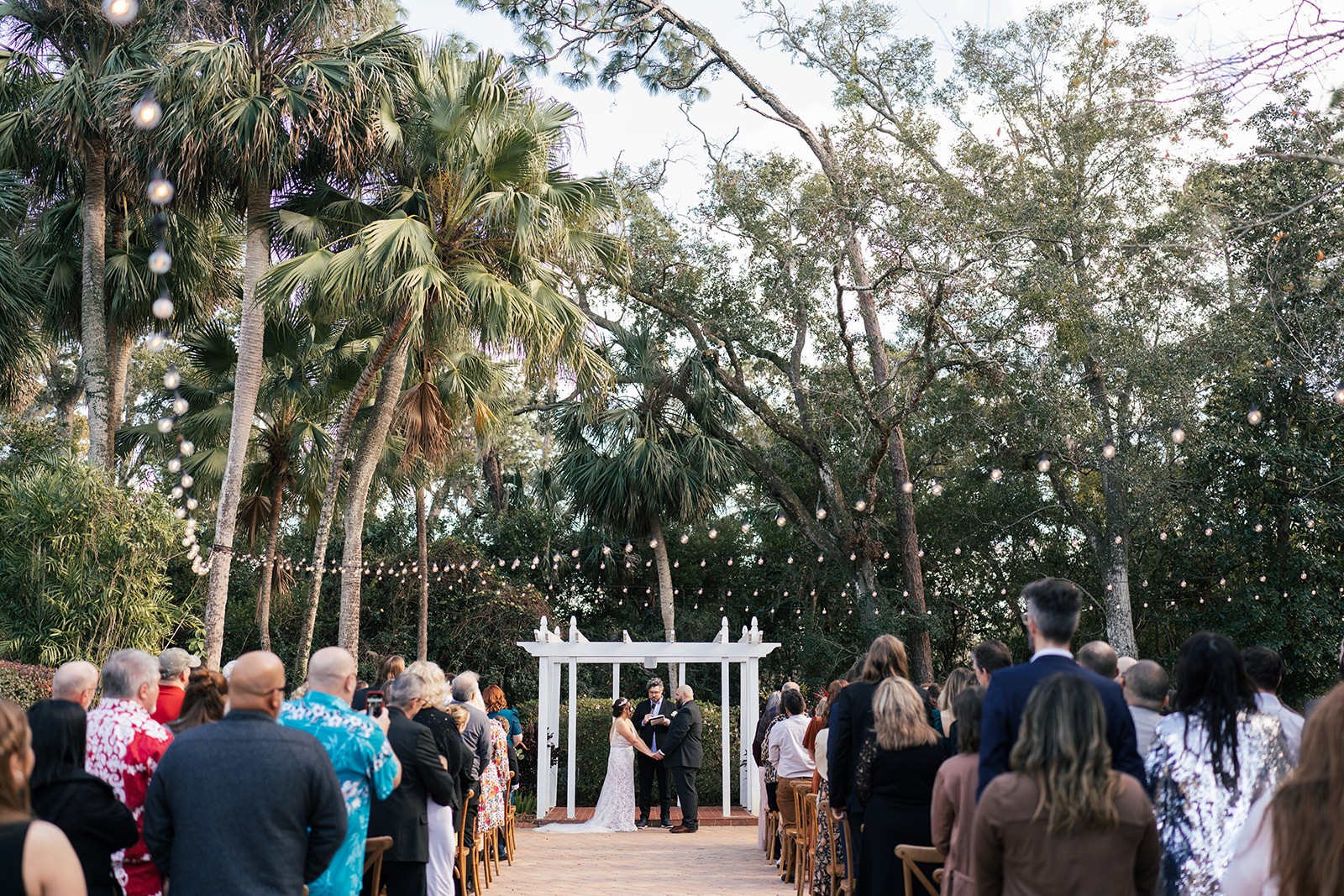 Wide view of the couple at the altar under string lights and tall palms as guests look on