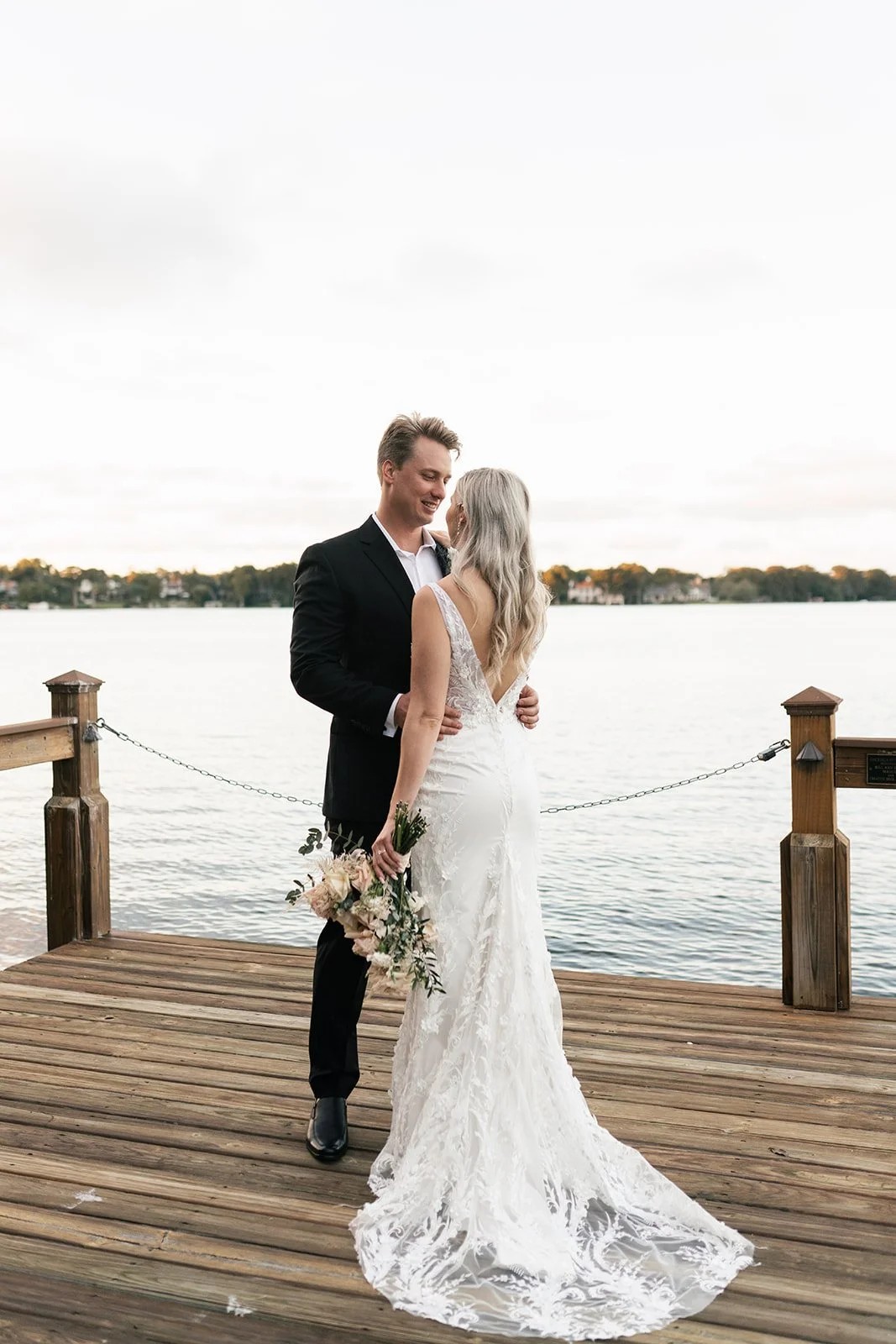 Bride and groom face each other on wooden dock above still water