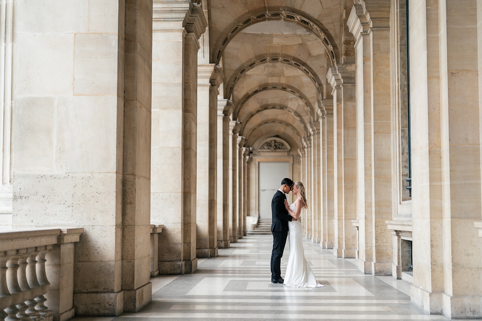 Bride and groom standing forehead to forehead in a grand stone colonnade with arched vaulting, a destination wedding portrait