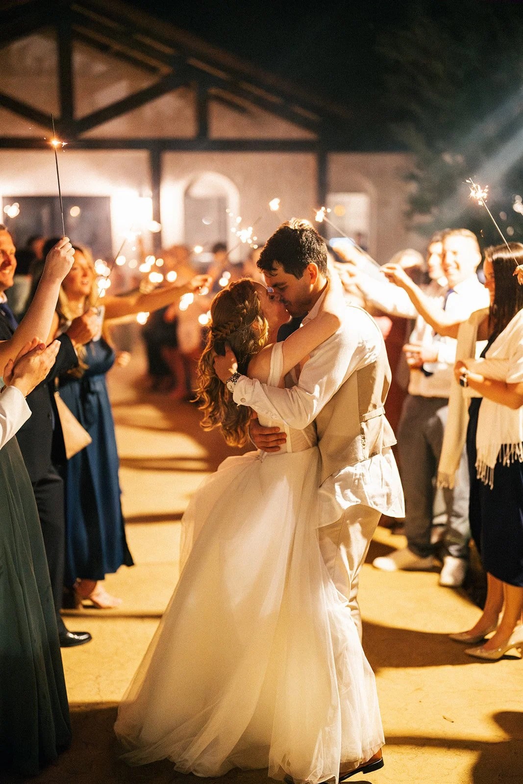 Bride and groom kiss surrounded by guests waving sparklers at night
