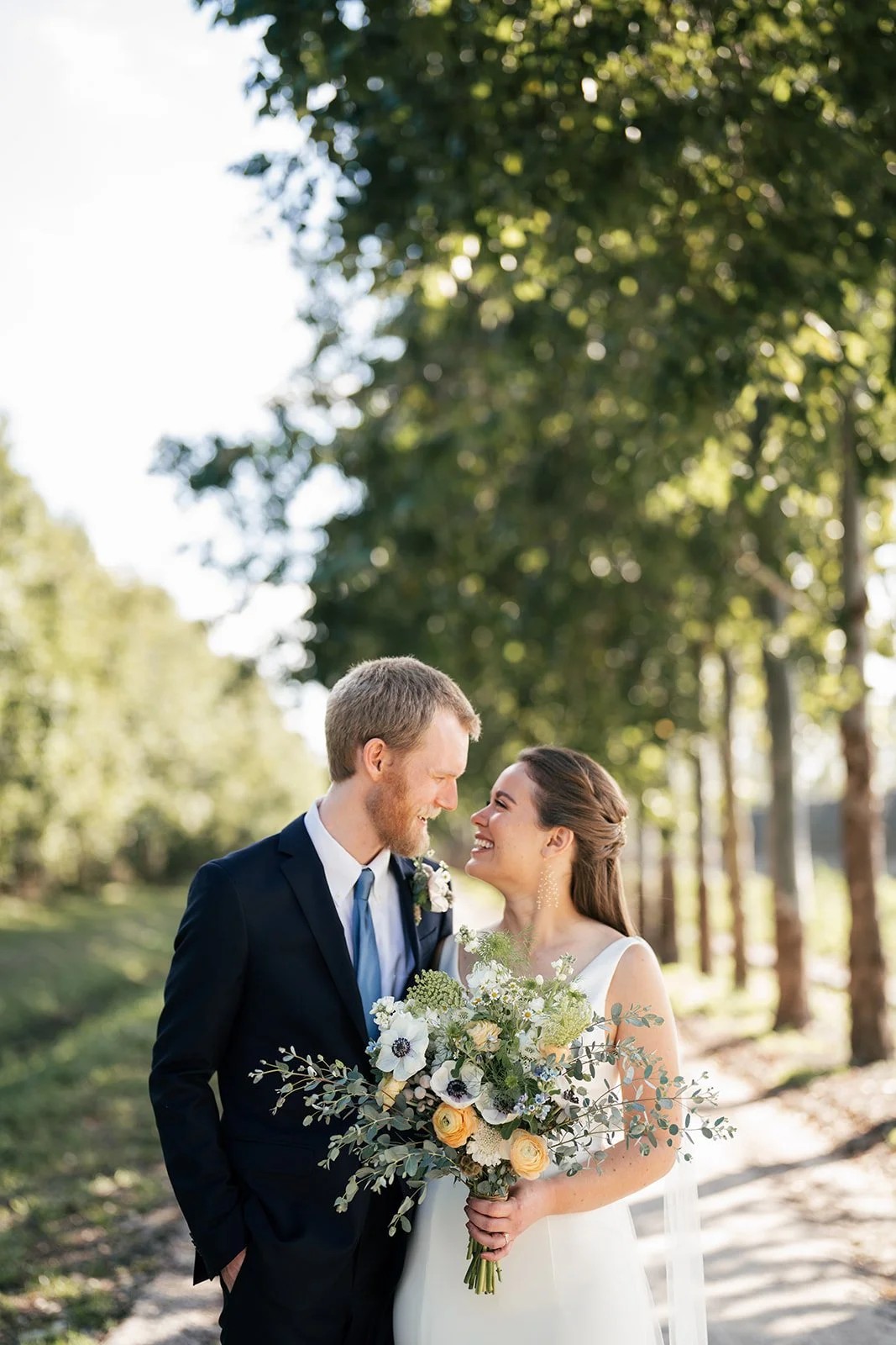 Bride smiles at groom while holding wildflower bouquet between trees