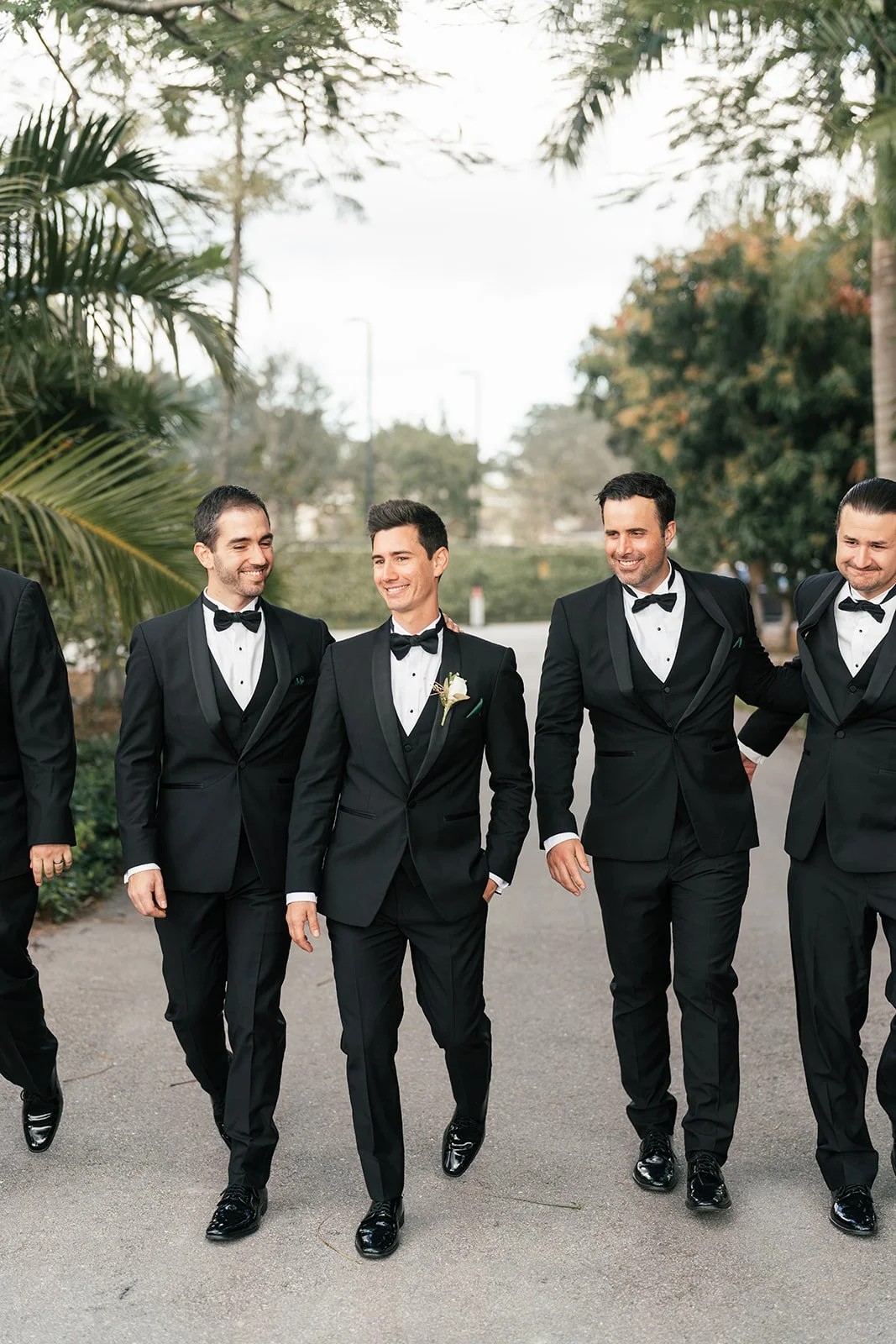 Groom walks with four groomsmen down a palm-lined drive in black tuxedos