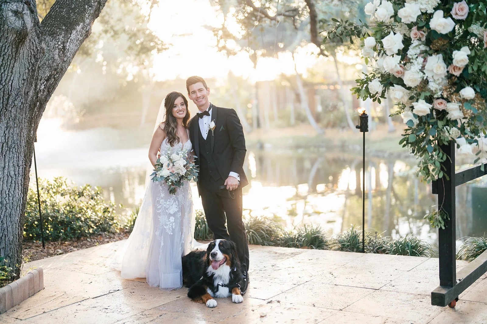 Bride, groom, and Bernese mountain dog pose by a pond at sunset