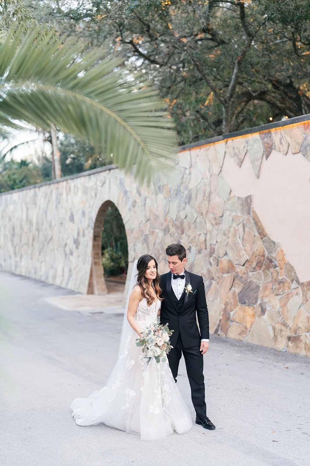 Bride and groom walk together along stone wall with palm frond overhead