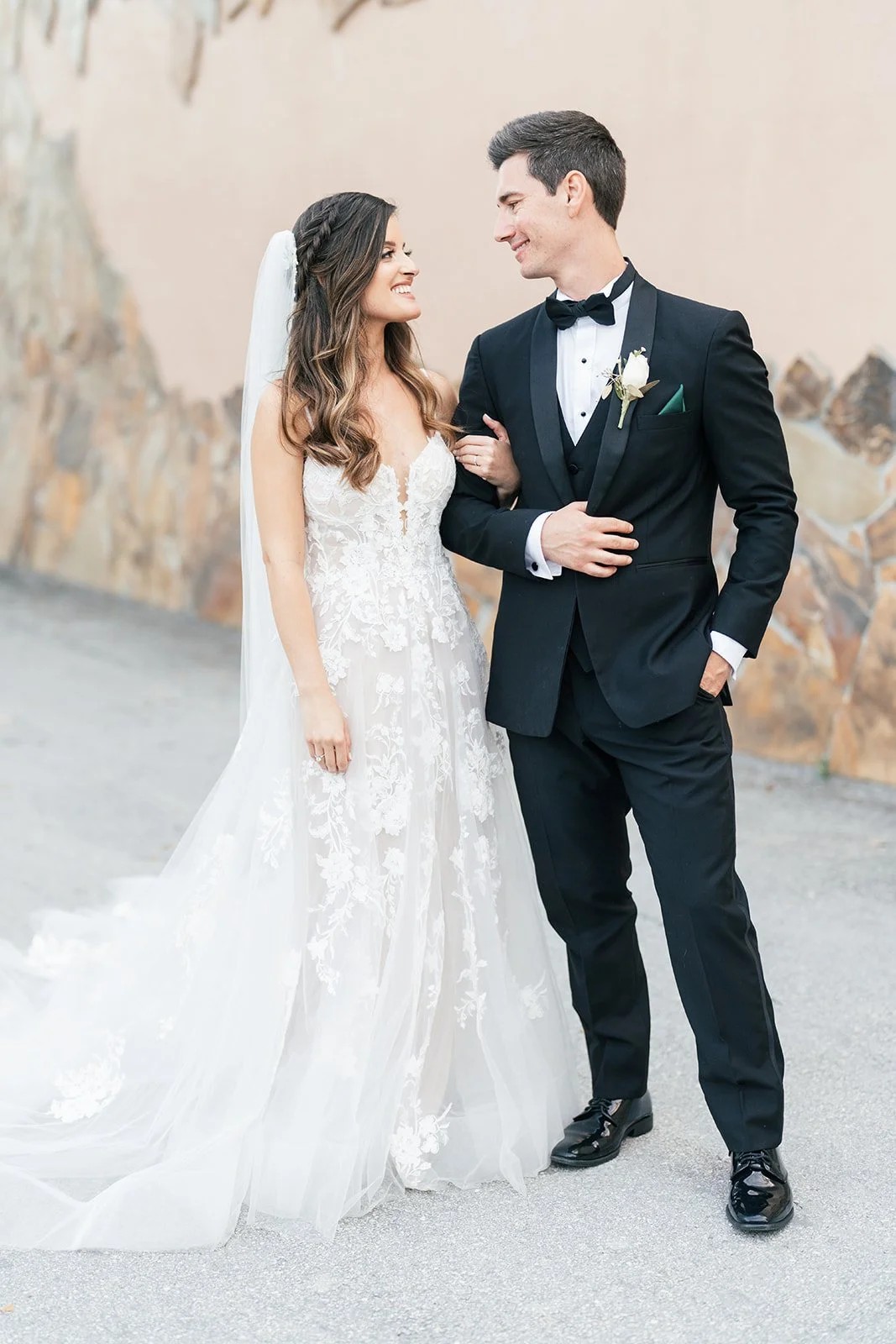 Bride in floral lace gown laughs with groom in black tuxedo outdoors