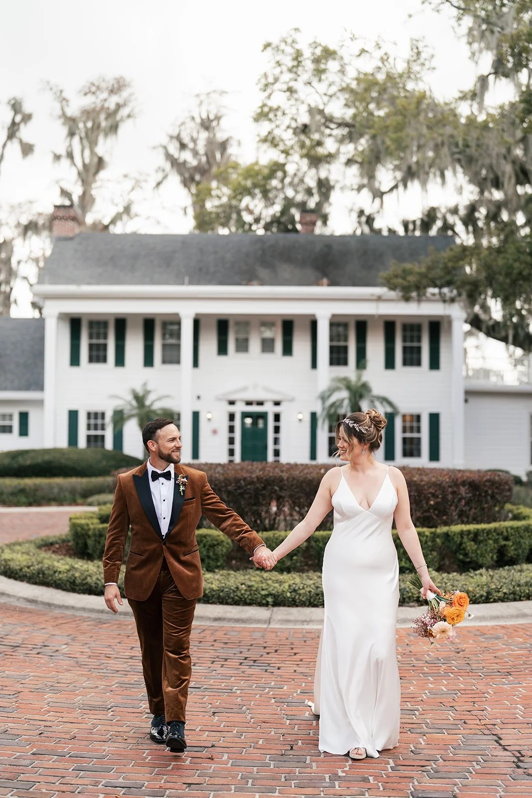 Couple holds hands on brick drive in front of large white colonial home