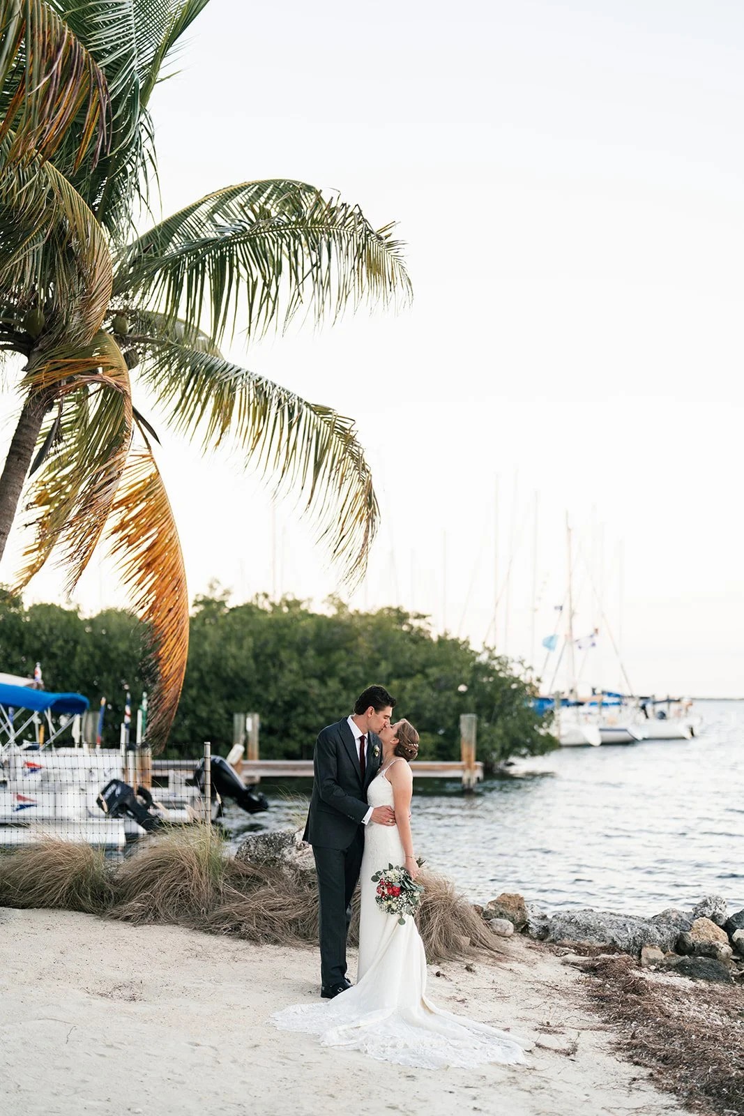 Bride and groom stand on sand beneath palm beside boats at dusk