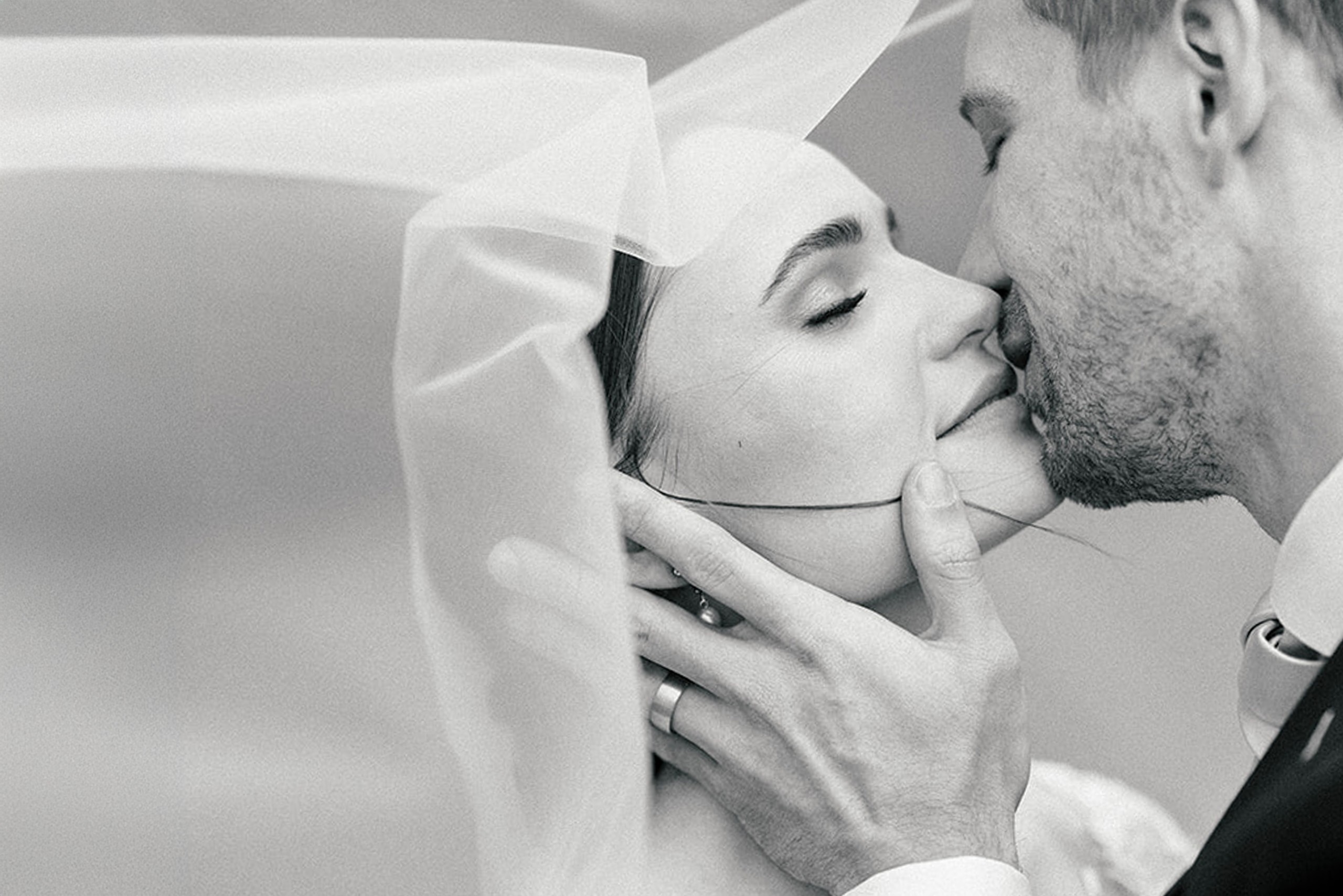 Black-and-white portrait of a bride and groom kissing beneath a wind-caught veil, soft bokeh behind them