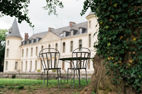 Two metal chairs sit at a small round table on grass directly beneath a towering old tree.