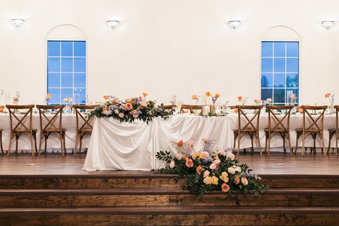 Empty reception space showing the decorated sweetheart table with abundant coral and pink floral centerpiece framed by tall blue-paned windows.