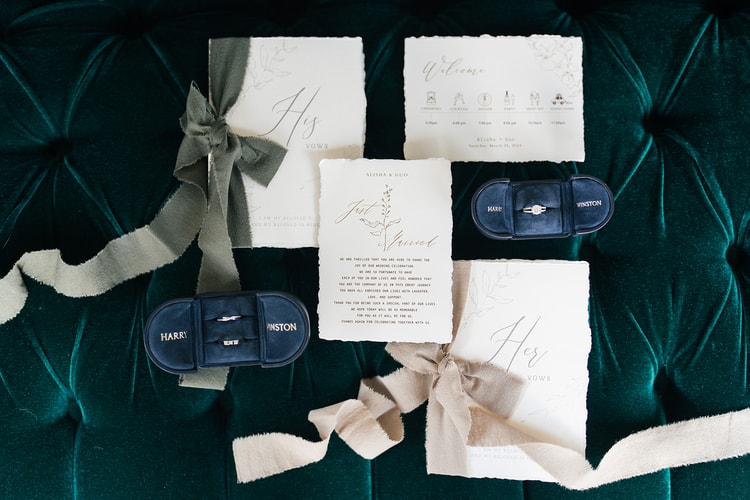 Flat-lay wedding stationery suite including vow booklets, a welcome card, rings in navy velvet boxes, and a schedule card arranged on a tufted emerald surface.
