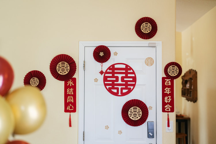 Door decorated with traditional Chinese double-happiness symbols, red paper fans, and wedding couplets in gold beside red and gold balloons for getting-ready festivities.