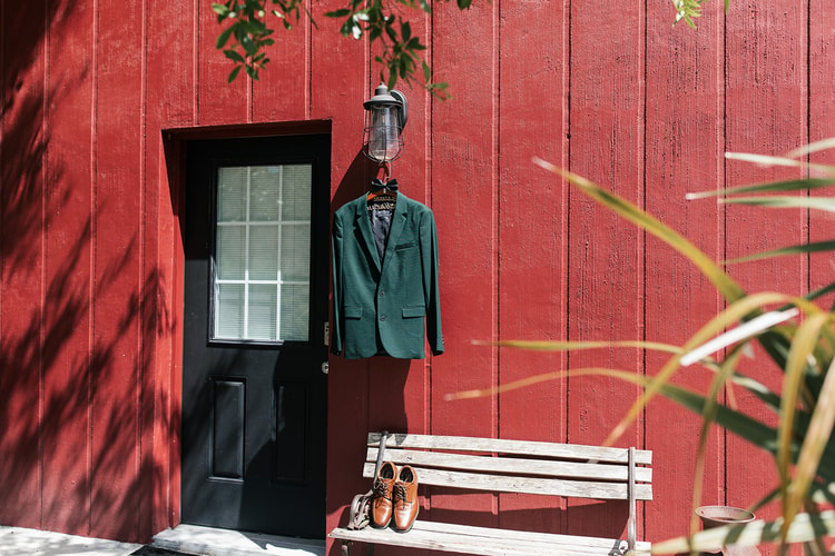 Groom's dark green suit jacket hanging on a lamp hook beside a red barn door, with brown dress shoes placed neatly on a wooden bench below.