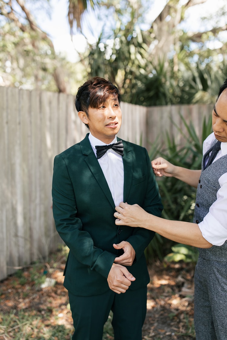 Groom in a forest-green tuxedo having his lapel adjusted by a groomsman outdoors, both looking focused as they get ready together.