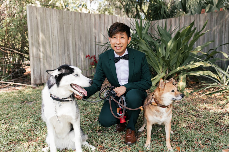 Groom in a green suit crouching between two dogs, a husky and a Shiba Inu, on the lawn before the ceremony, grinning broadly.