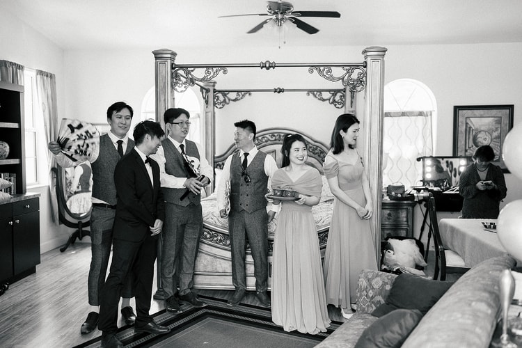 Black-and-white getting-ready scene with groomsmen and bridesmaids gathered in a bedroom, chatting and laughing ahead of the ceremony.