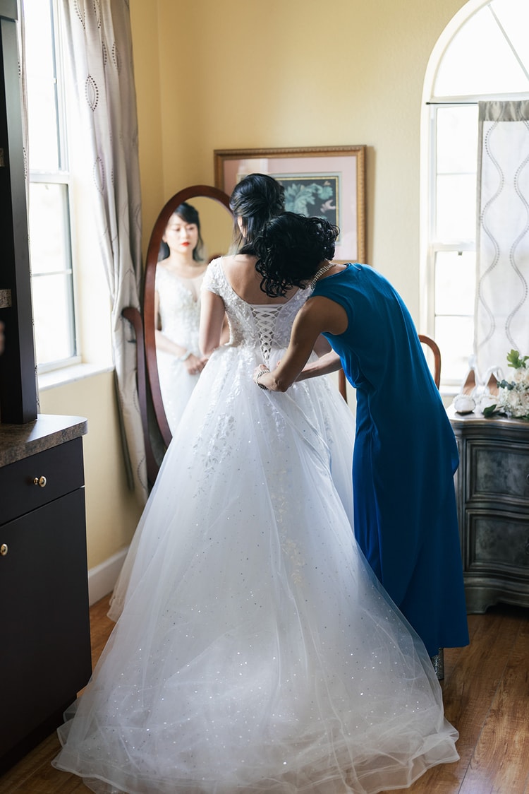 Getting ready: a woman in a blue dress laces the back of the bride's ball gown while the bride faces a floor mirror, her reflection visible in the glass.