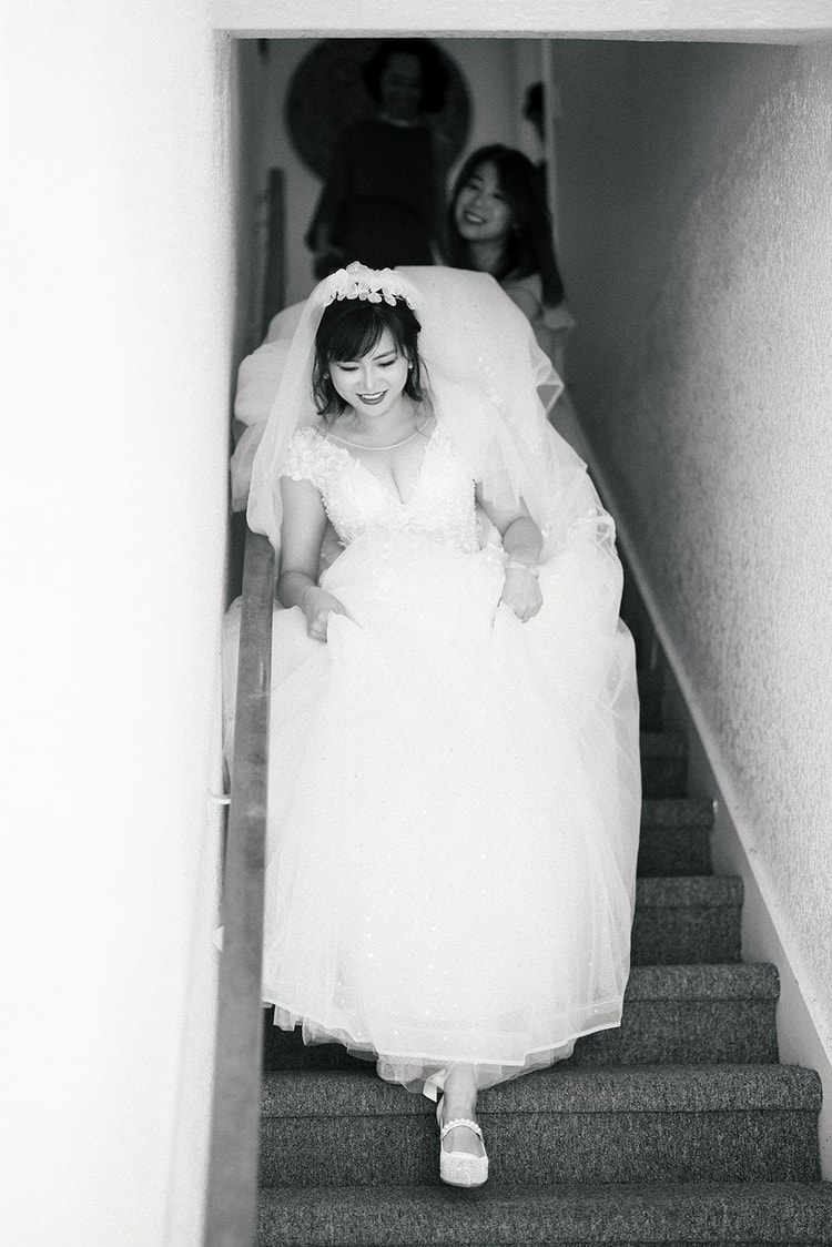 Black-and-white portrait of the bride descending a staircase in her ball gown and veil, smiling down, with a bridesmaid visible watching from above.