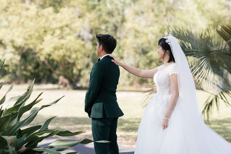 First look: the bride reaches forward to tap the groom's shoulder from behind as he stands facing away, her expression full of anticipation under palm trees.