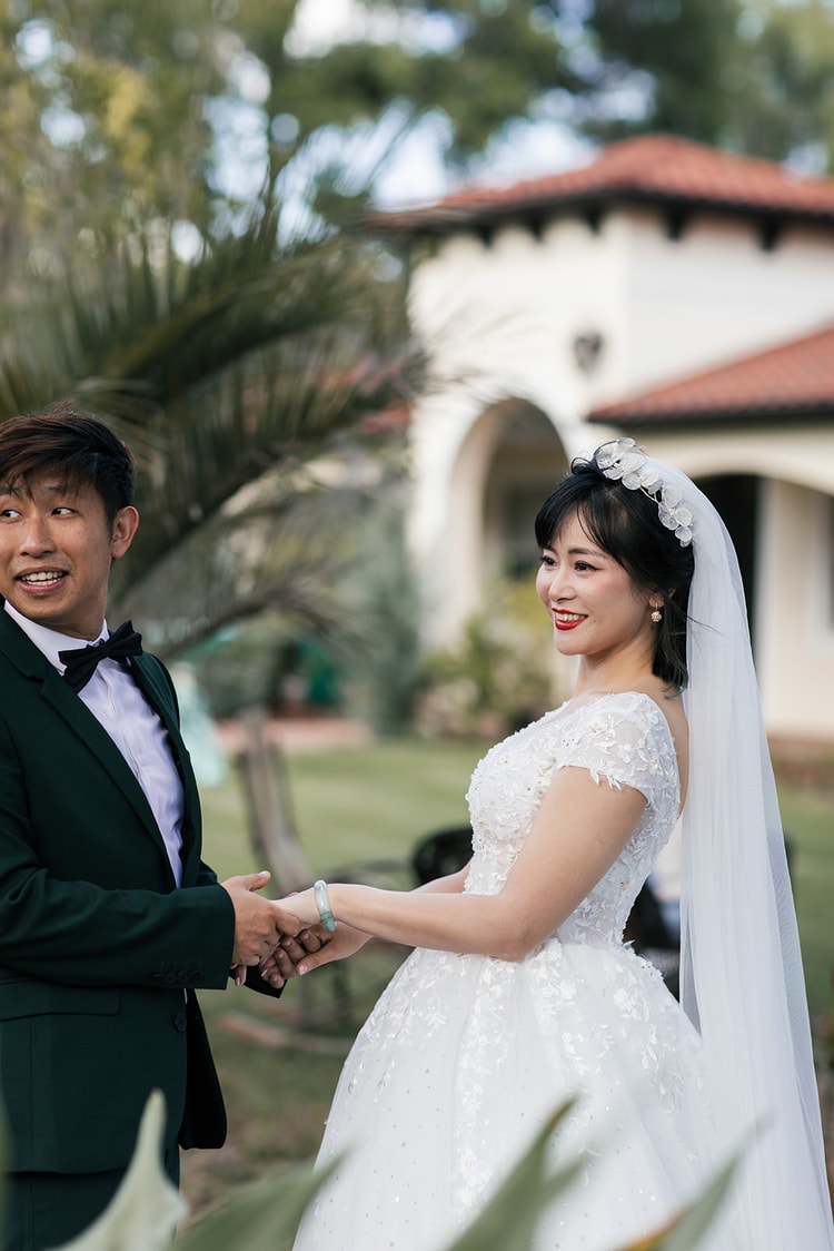 Couple holding hands and laughing during portraits outdoors, a Mediterranean-style building with a red-tile roof softly blurred in the background.