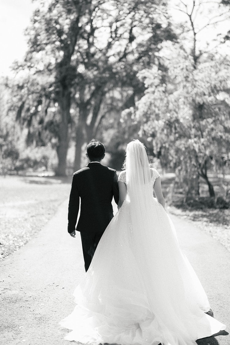 Black-and-white portrait of the couple walking away down a tree-lined path, the bride's full cathedral train trailing behind her.
