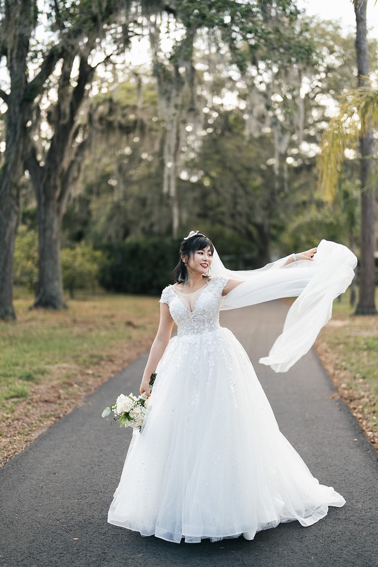 Bride in a lace ball gown twirling her veil on a shaded garden path lined with moss-draped oaks, bouquet in hand, looking joyful.