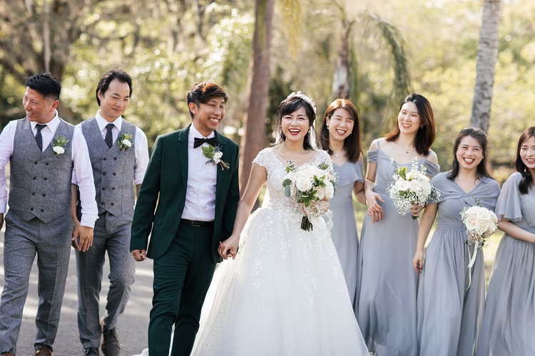 Full wedding party walking together in bright sun, the bride and groom at the center holding hands while bridesmaids in gray-blue and groomsmen in gray vests flank them.