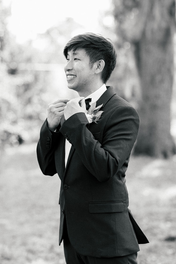 Black-and-white close-up of the groom adjusting his bow tie before the ceremony, smiling to himself with a boutonniere on his lapel.