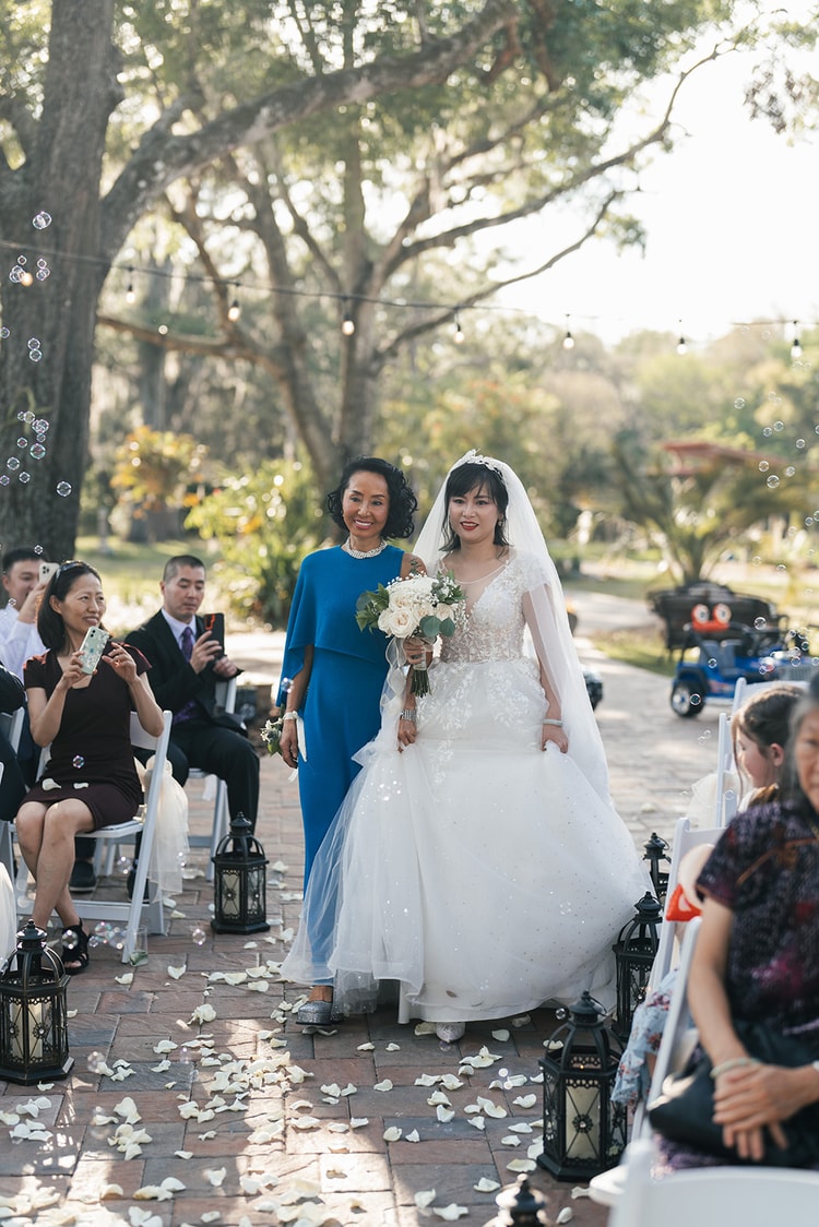 Processional: bride walks the petal-strewn aisle escorted by a woman in blue, guests watching from white chairs on either side as soap bubbles drift through the air.