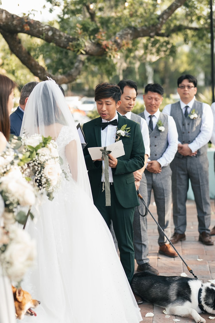 Groom reads his vows from a card at the outdoor ceremony altar, groomsmen standing behind him, the couple's dog leashed nearby on a brick patio.