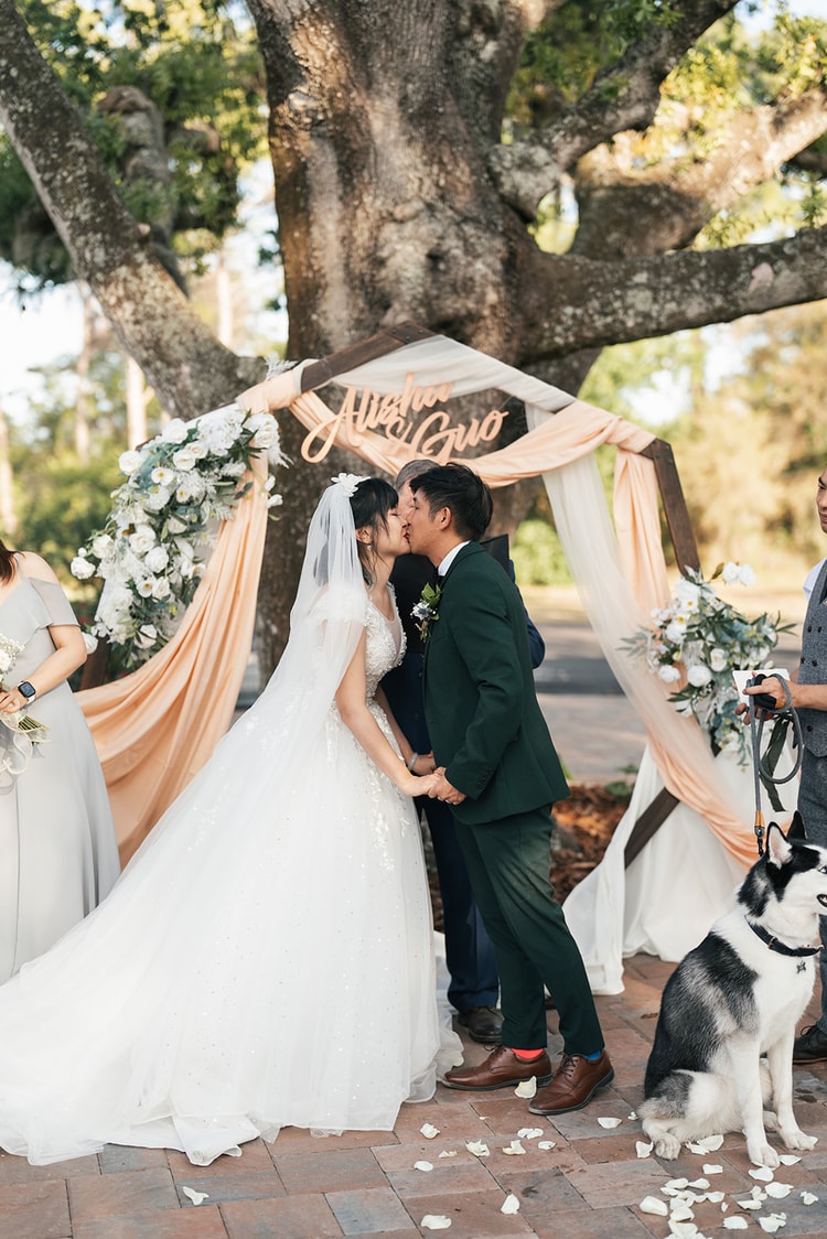 First kiss under a wood arch draped in peach fabric and white florals, with a laser-cut name sign reading Alisha and Guo overhead and their husky at their side.