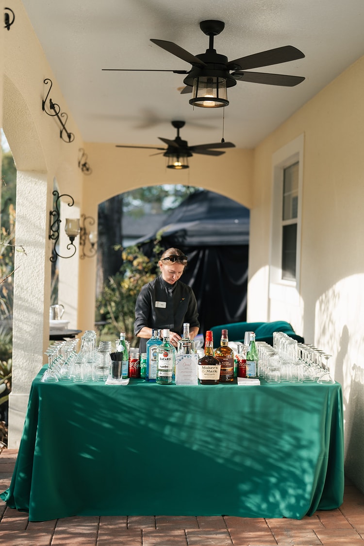 Bartender setting up the outdoor cocktail-hour bar, bottles of spirits and rows of glassware arranged on a teal-draped table under a covered porch.