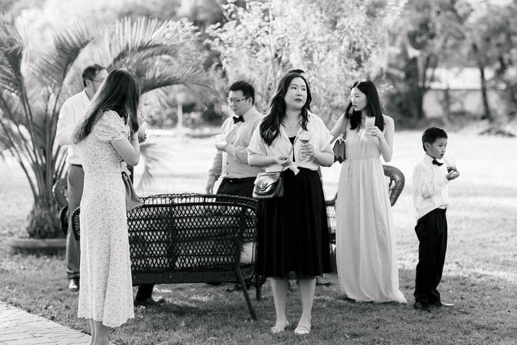 Black-and-white candid of guests mingling on the lawn during cocktail hour, drinks in hand, a young boy in a bow tie joining the group.