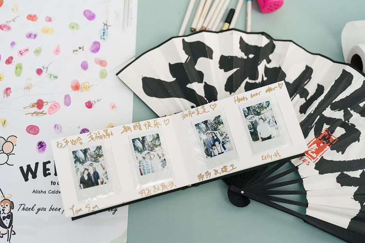 Reception guest-book table displaying a fingerprint tree, Instax photo strip booklet with handwritten messages in English and Chinese, and a calligraphed paper fan.
