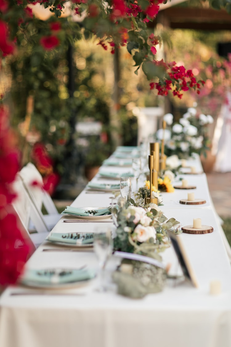 Head table detail: long white linen table set with sage napkins, gold taper candles, a greenery and rose garland runner, beneath a canopy of red bougainvillea.