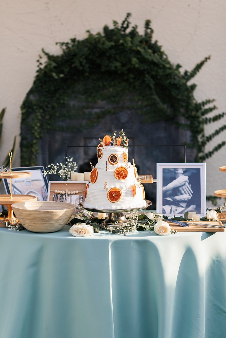 Three-tier white wedding cake decorated with dried orange slices displayed on a blue-clothed dessert table with white roses, greenery, and framed photos.