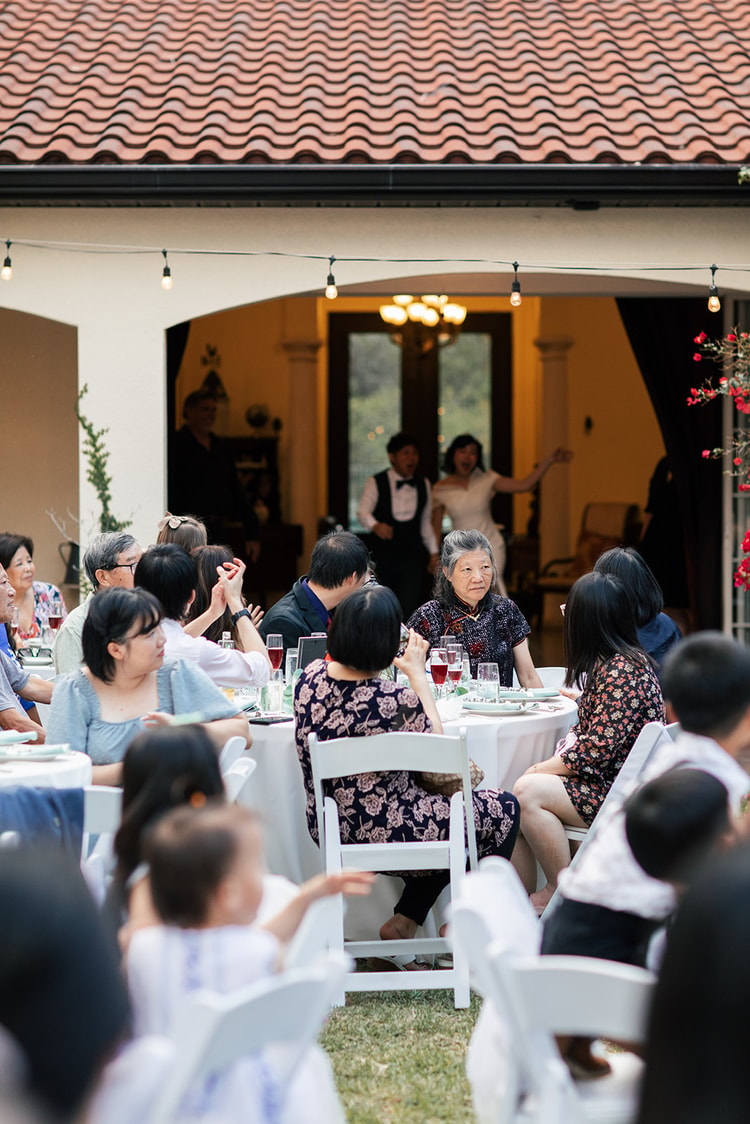 Reception in full swing: guests seated at round tables applaud as the bride and groom make an entrance through arched doors glowing with warm interior light.
