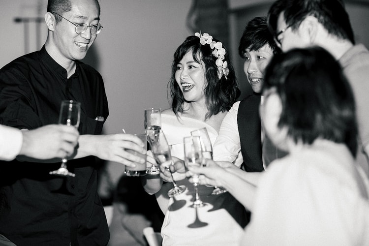 Black-and-white reception candid: the bride in a white off-shoulder dress laughs as she clinks champagne flutes with guests gathered close around her.