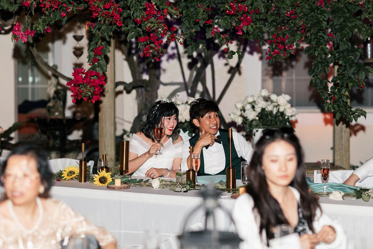Couple seated at the sweetheart table during reception speeches, sharing a relaxed laugh with champagne in hand, sunflowers and white roses on the table.