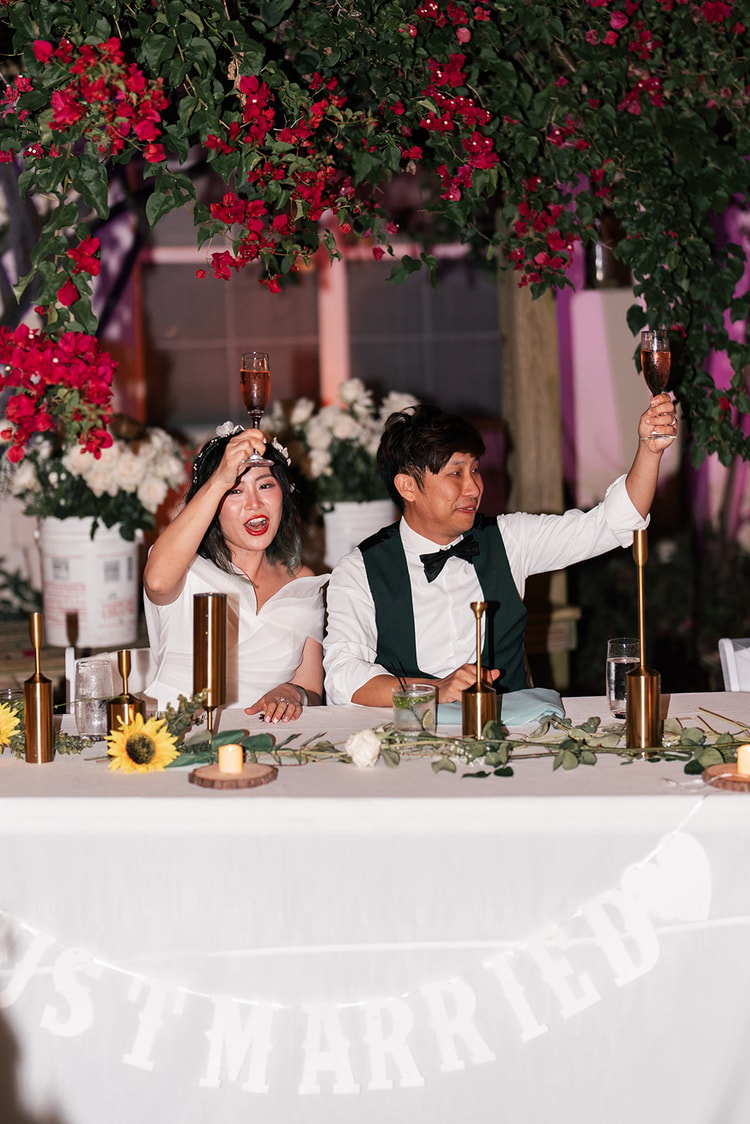 Bride and groom raise their champagne flutes from the sweetheart table at the outdoor reception, a Just Married banner below and red bougainvillea behind them.