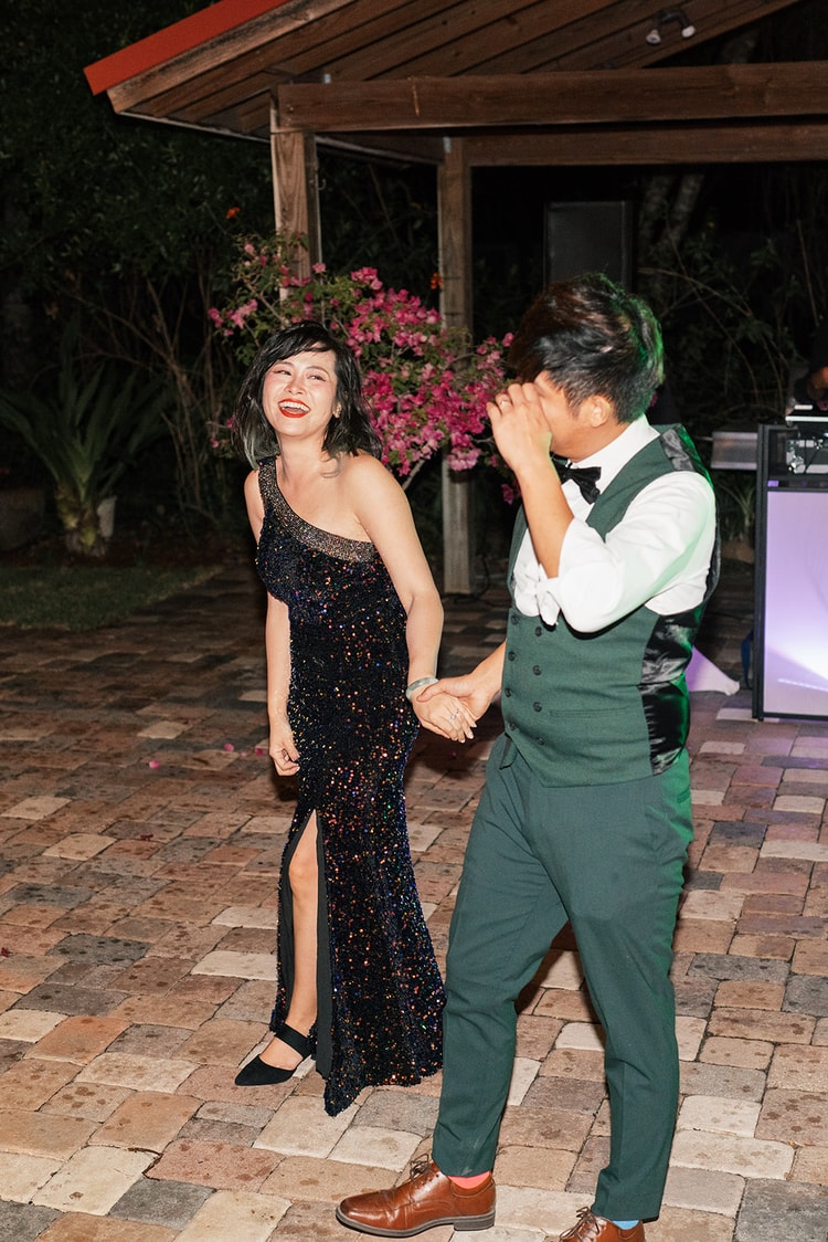 Bride and groom dancing together outdoors at night, both laughing freely, the groom covering his face in delight as bougainvillea blooms behind them.
