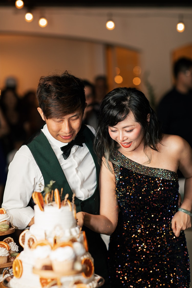Cake cutting: the couple leans over their citrus-adorned wedding cake together, both in their reception outfits, laughing as they prepare to cut the first slice.