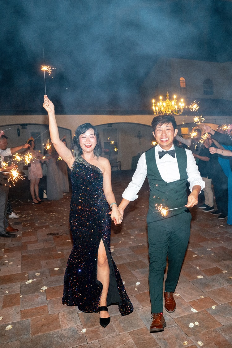 Couple walking hand-in-hand through a sparkler tunnel at the end of the night, both holding sparklers high, smiling radiantly in their reception outfits.