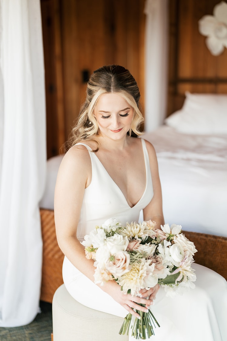 Bride in a slip-style gown seated on an ottoman, gazing down at her lush white and blush bouquet of dahlias and ranunculus.