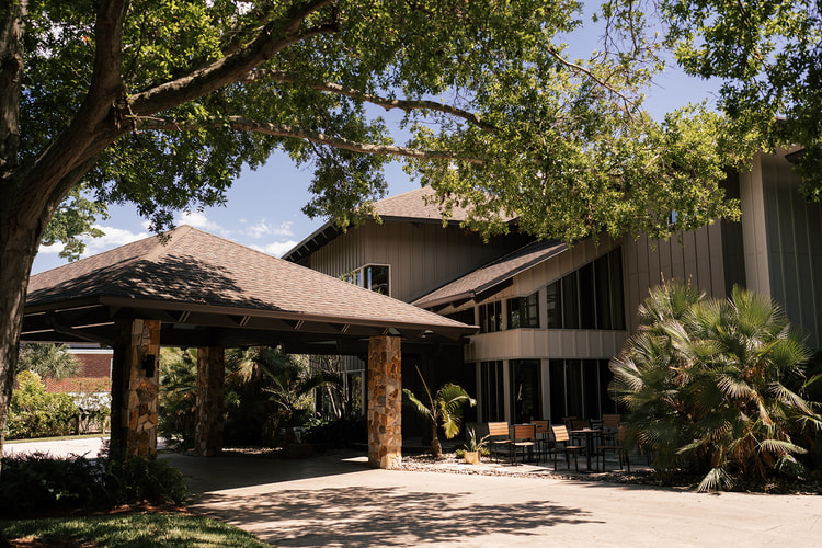 Exterior of a wood-and-stone venue building shaded by large trees on a sunny afternoon.