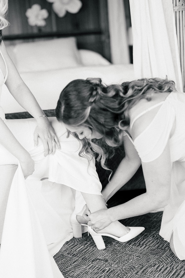 Black-and-white getting-ready moment: a bridesmaid kneels to fasten the bride's white heels while another steadies her dress.